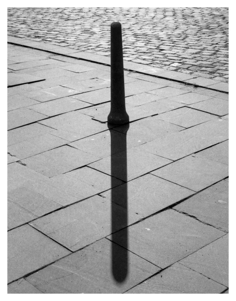 Ein Straßenpoller auf einem gefliesten Untergrund, im Hintergrund Kopfsteinpflaster. Der Poller wirft einen Schatten, der gerade nach unten zeigt.