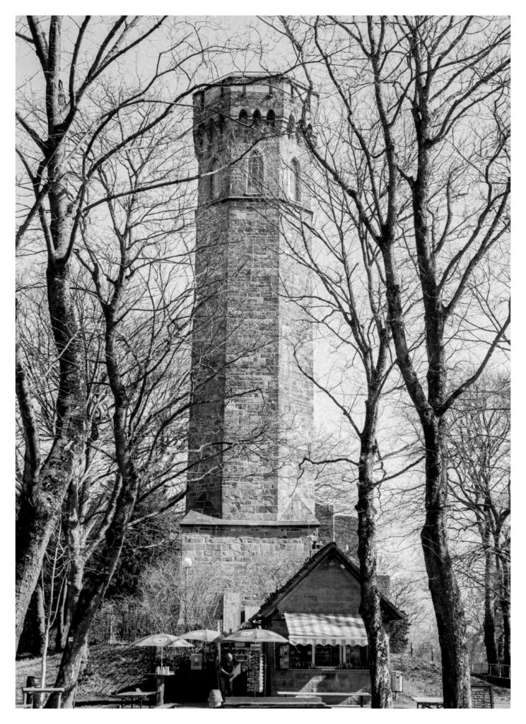 Altertümlicher Turm zwischen Bäumen. Am Fuße des Turms ist ein Kiosk mit Sonnenschirmen.